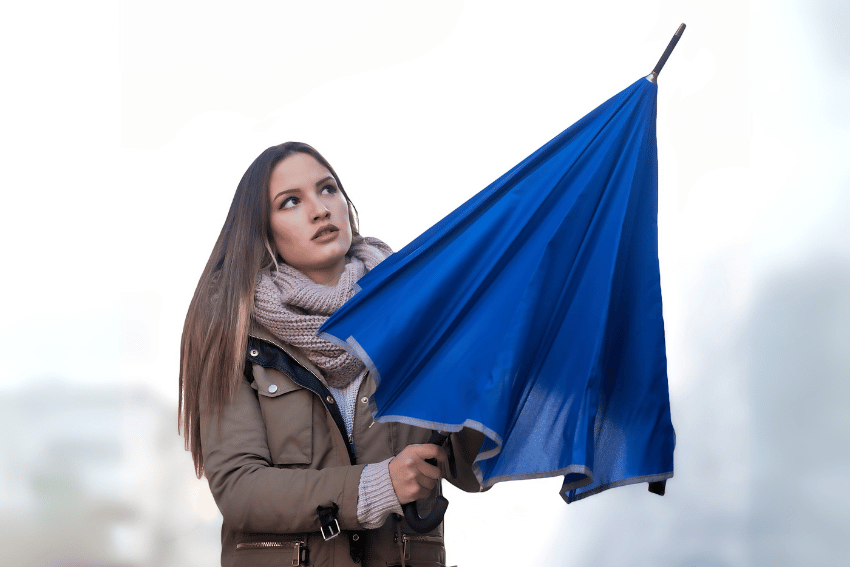 A young woman wearing a scarf and coat holds a blue umbrella turned inside out by the wind. She looks up with a concerned expression, as if witnessing how to break up a dog fight. The background is blurred and outdoors.