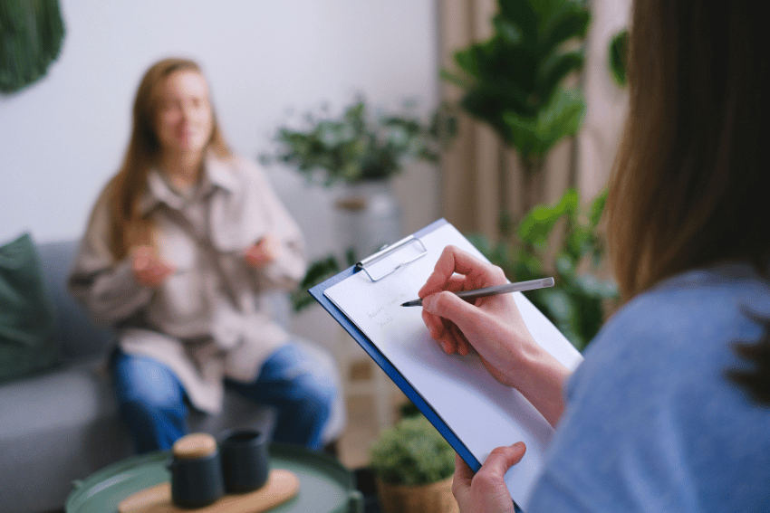 A person holds a clipboard and writes while facing another person sitting on a couch, gesturing with their hands, in a room with plants and a small table—discussing Emergency Pet Boarding NDIS options.