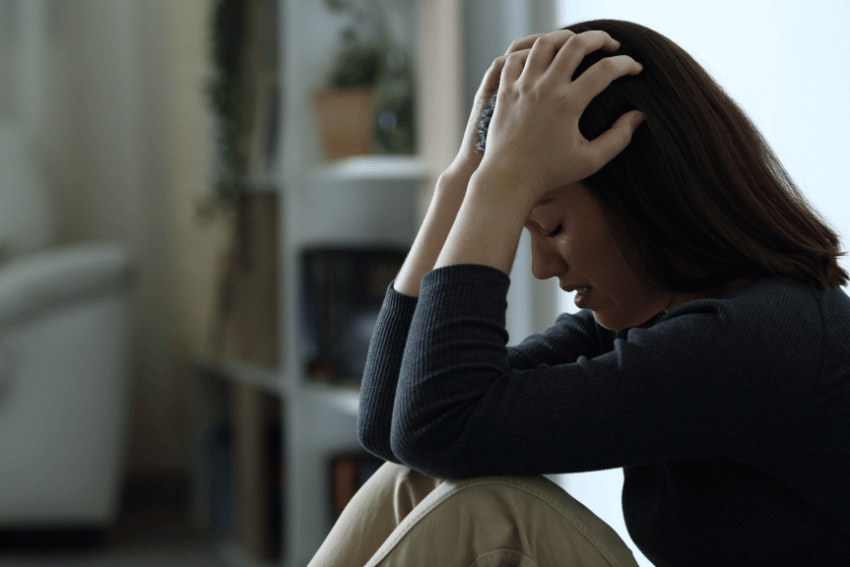 A woman sits on the floor indoors, looking stressed as she leans forward with her elbows on her knees and hands gripping her head—possibly worried about finding Emergency Pet Boarding NDIS support.