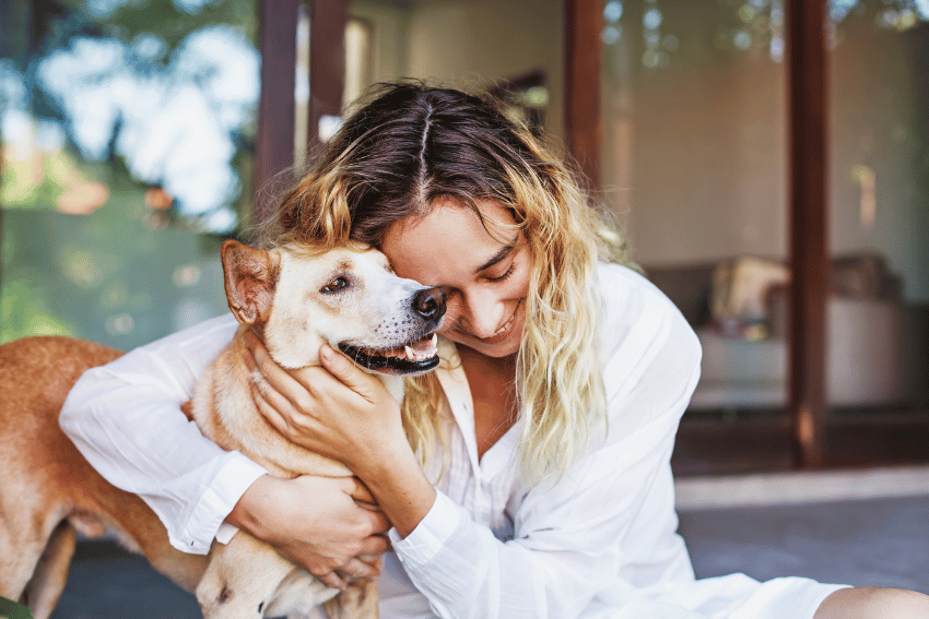 A woman in a white shirt hugs a happy tan dog outside a house, both appearing joyful and content—showcasing the comfort Emergency Pet Boarding NDIS can bring to pet owners in need.